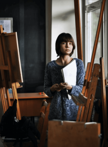 female standing with art book next to a wooden easel
